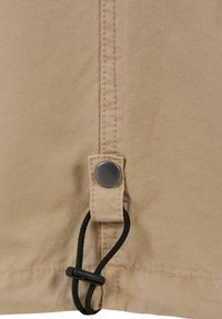 Section de tissu beige avec une couture verticale, un bouton-pression métallique et un cordon élastique noir passé dans une languette en tissu.