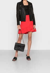 Zwarte leren motorjack, zwart geknoopt topje, rode gerimpelde rok, zwarte geweven slippers en een zwarte handtas met krokodillenprint en kettingband.