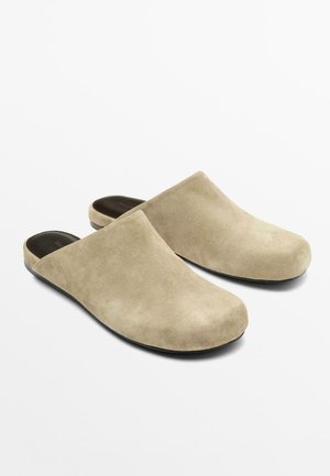 Paire de chaussons beige en suède à enfiler, avec des bouts fermés et des semelles intérieures noires rembourrées sur fond blanc.