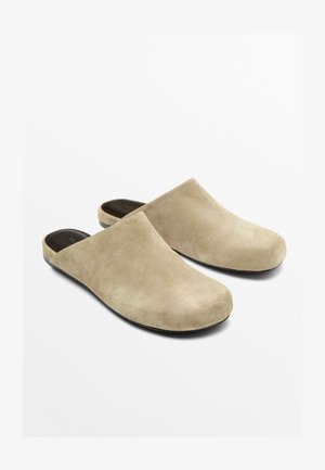 Paire de chaussons beige en suède à enfiler, avec des bouts fermés et des semelles intérieures noires rembourrées sur fond blanc.
