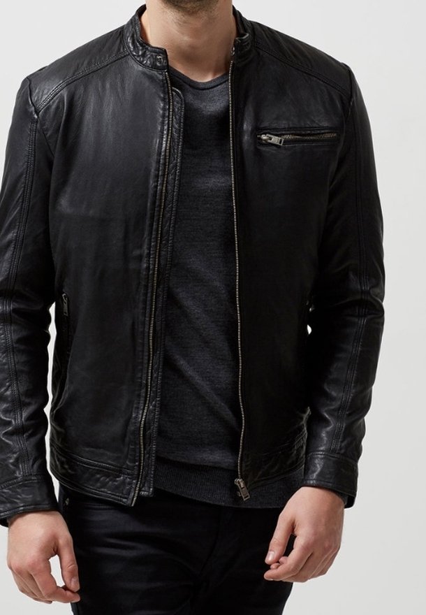 Homme portant une veste en cuir noire sur une chemise gris foncé, les bras détendus le long du corps, sur un fond clair uni.
