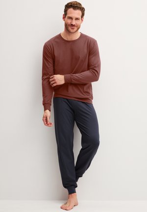 Man staat op blote voeten, draagt een bruin shirt met lange mouwen en donkerblauwe joggingbroek tegen een effen witte achtergrond.