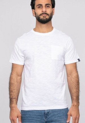 Homme barbu portant un T-shirt blanc à manches courtes et poche poitrine, debout devant un fond gris clair uni.