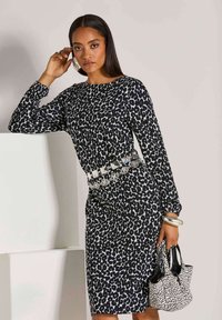 Frau in einem schwarz-weißen Leopardenmuster-Kleid mit silbernem Gürtel, die eine passende gepunktete Handtasche hält, große Creolen und Armbänder trägt.