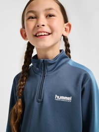 Smilende barn med langt brunt fletning har en blå Hummel Performance lynlås sportsjakke på mod en ensfarvet baggrund.