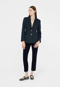Marineblauer Blazer mit strukturiertem Muster, Spitzrevers, zwei Knöpfen und Vordertaschen. Kombiniert mit eng anliegenden dunklen Hosen und grauen Ballerinas.