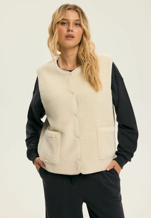 Kremowa kamizelka z polaru z okrągłym dekoltem, guzikami z przodu i dwoma naszywanymi kieszeniami. Noszona na czarnej bluzce z długim rękawem i dopasowanych spodniach.