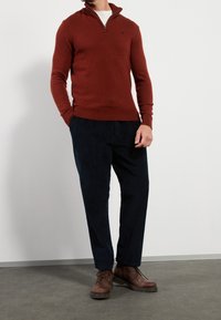 Roestkleurige gebreide trui met een ritskraag, gecombineerd met marineblauwe ribfluwelen broek en bruine veterschoenen. Eenvoudig ontwerp, gestructureerd breisel.