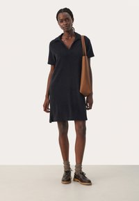 Abito polo blu navy con maniche corte e colletto, presenta spacchi laterali. Abbinato a una borsa in pelle marrone e scarpe marroni con lacci e suola spessa.