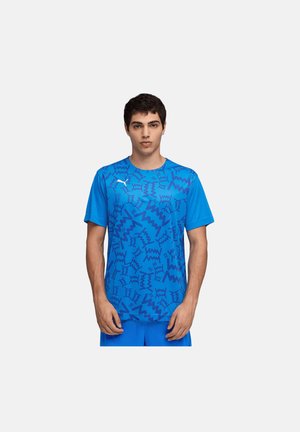 Junger Mann trägt ein blaues Puma-Sportshirt mit abstrakten dunkelblauen Zickzackmustern und steht vor einem einfarbigen hellen Hintergrund.