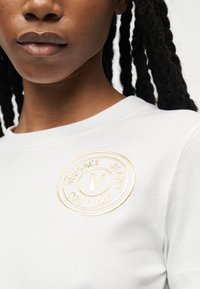 Κοντινό πλάνο ενός ατόμου που φοράει λευκό πουκάμισο με χρυσό λογότυπο Versace Jeans Couture στο στήθος, με εμφανή τα χείλη και μέρος πλεξούδας μαλλιών.