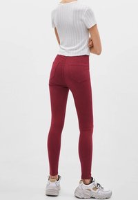 Mujer de pie con los brazos cruzados, vistiendo una camiseta blanca de manga corta de canalé, jeans skinny burdeos y zapatillas chunky blancas sobre un fondo liso.