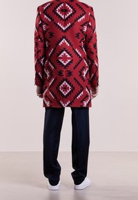 Manteau rouge à motifs géométriques en noir et blanc, associé à un pantalon bleu marine foncé et des baskets blanches.