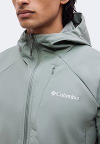 Άνδρας που φοράει ανοιχτό γκρι μπουφάν Columbia με κουκούλα, εμπρόσθιο φερμουάρ και μικρή τσέπη με φερμουάρ στο στήθος, σε απλό φόντο.