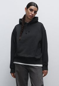 Felpa nera con una vestibilità rilassata, cappuccio con cordino e polsini a costine, indossata sopra una maglietta bianca. Abbinata a jeans grigi, design minimal.