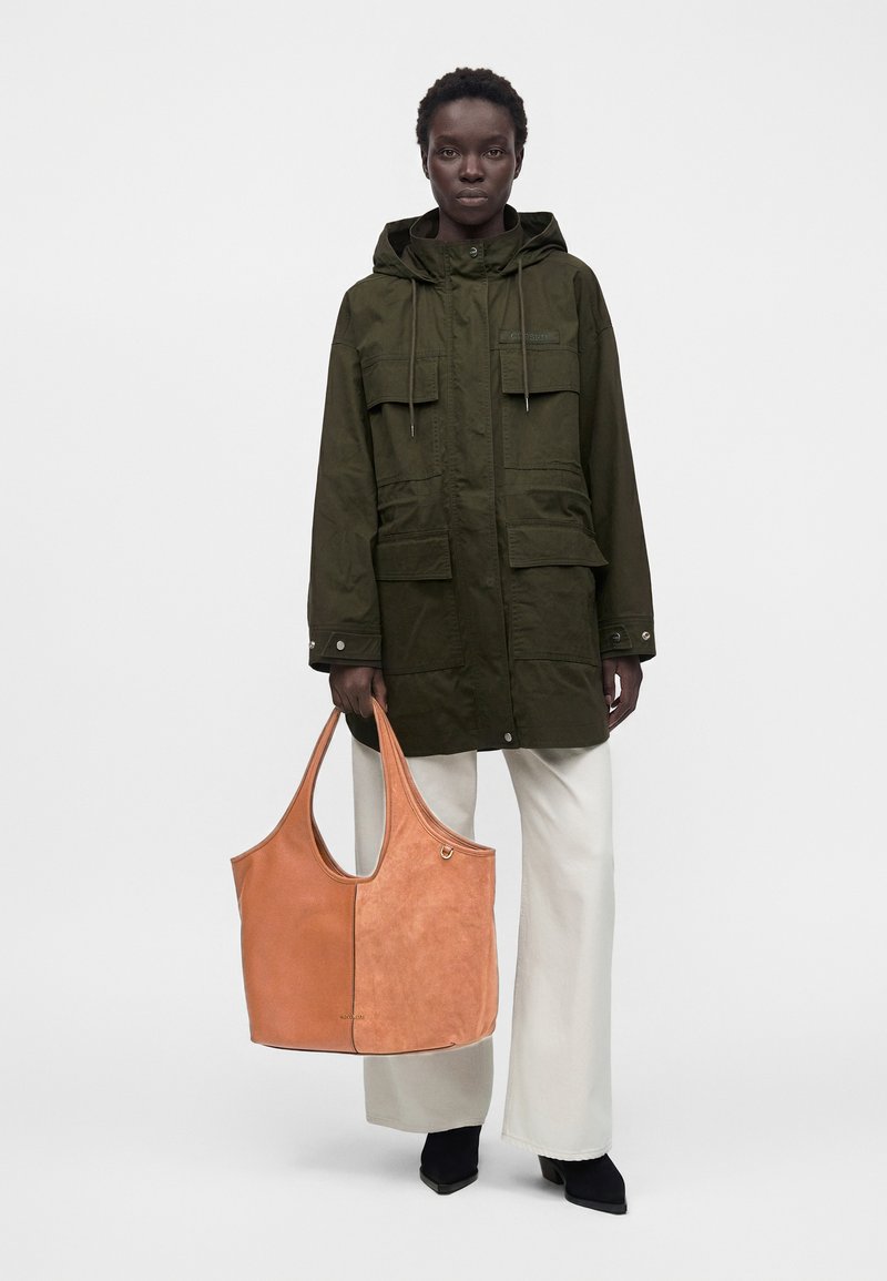 Femme portant une veste à capuche vert olive, pantalon blanc à jambes larges, chaussures noires pointues, tenant un grand sac cabas en cuir marron clair.