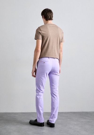 Camiseta castanha de manga curta combinada com calças lilás ajustadas. As calças têm bolsos traseiros e são feitas de um tecido suave. Sapatos pretos.