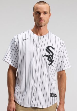 Man draagt een wit Chicago White Sox honkbalshirt met zwarte dunne strepen en zwarte logo's, staand voor een neutrale grijze achtergrond.
