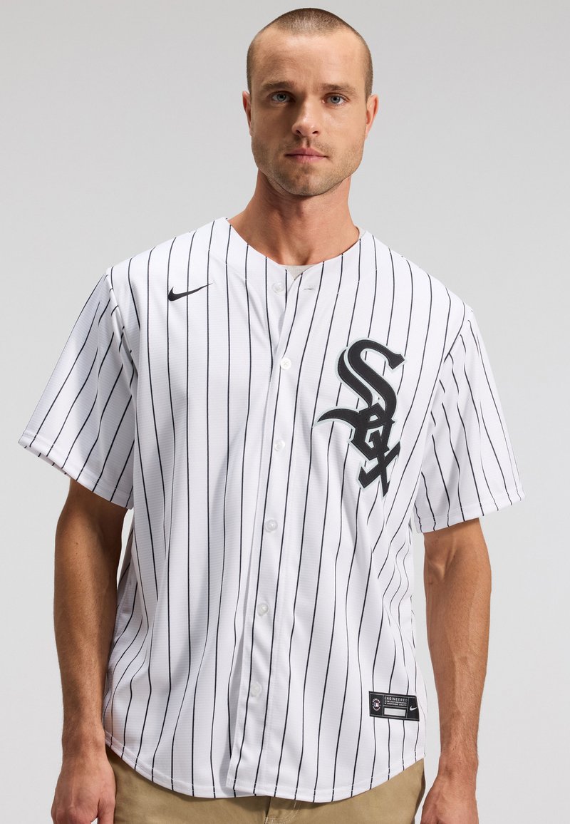 Άνδρας που φοράει λευκή μπλούζα baseball Chicago White Sox με μαύρες λεπτές ρίγες και μαύρα λογότυπα, στέκεται μπροστά σε ουδέτερο γκρι φόντο.