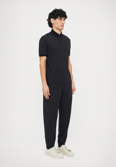 Μαύρο πόλο μπλουζάκι με υφή υφάσματος, κοντά μανίκια και γιακά, συνδυασμένο με μαύρο παντελόνι φόρμας και ανοιχτόχρωμα �αθλητικά παπούτσια.