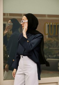 Granatowa koszula z długim rękawem z dużym guzikiem, biały spódnicowy materiał w wysokim stanie, czarny hijab i białe okulary przeciwsłoneczne. Lustrzana powierzchnia w tle.
