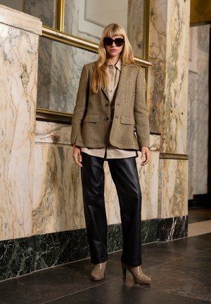 Blazer en pied-de-poule marron, chemise crème à boutons, pantalon en cuir noir et escarpins en imprimé serpent. Miroir à cadre doré en arrière-plan.