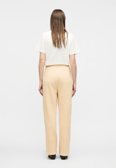 Γυναίκα με μακριά ίσια μαλλιά, που φοράει λευκό τοπ με κοντά μανίκια και μπεζ παντελόνια φαρδιάς γραμμής, στέκεται γυρισμένη προς τα πίσω σε απλό φόντο.