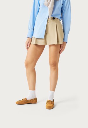 Donna che indossa pantaloncini beige a pieghe, camicia azzurro chiaro con bottoni, calzini bianchi e scarpe in stile mocassino color cuoio, in piedi davanti a uno sfondo neutro.
