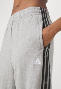 Close-up of a person wearing grey Adidas sweatpants with black side stripes and a hand placed inside the pocket.