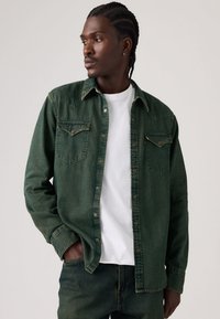 Πράσινο πουκάμισο από ντένιμ με δύο τσέπες στο στήθος, μυτερό γιακά και εμπρόσθιο κούμπωμα με κουμπιά. Φορεμένο πάνω από ένα λευκό T-shirt με άνετη εφαρμογή και μακριά μανίκια.