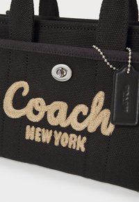 Crni platneni torba s izvezenim bež tekstom "Coach" i "New York", srebrnim hardverom i crnom oznakom s lancem.