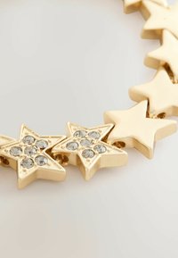 Aukso spalvos kaklo vėrinys, kuriame yra tarpusavyje sujungti žvaigždžių formų elementai, kai kurie iš jų puošiami skaidriais rhinestones, kad suteiktų papildomą detalumą ir tekstūrą.