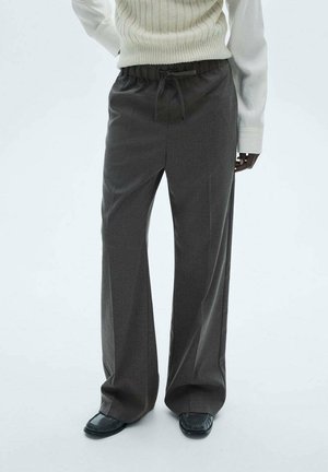 Personne portant un pantalon large gris foncé avec une taille à cordon, une chemise blanche à manches longues et des mocassins noirs sur un fond uni.