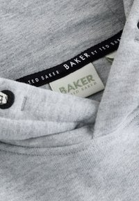Szary materiał w zbliżeniu, pokazujący obszar kołnierza z czarno-białymi metkami marki Ted Baker przyszytymi wewnątrz ubrania.