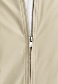 Tissu beige avec une fermeture éclair argentée partiellement ouverte, révélant un matériau blanc en dessous. Le curseur de la fermeture présente un design géométrique subtil.