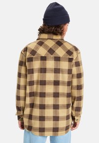 Beige-braun karierte Hemd aus Flanell mit abgerundetem Saum, Knopfmanschetten und einem Yoke-Detail. Getragen mit einer marineblauen Strickmütze.