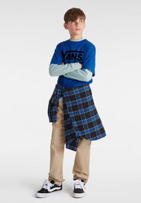 Sininen graafinen t-paita, jossa on "VANS"-logo, päällystetty vaaleansinisillä pitkillä hihoilla. Vaaleat housut, musta-valkoiset lenkkitossut ja sininen ruudullinen paita sidottuna vyötärölle.