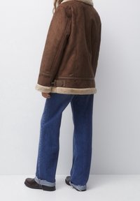Person iført brun shearlingjakke, blå opsmøgede vide jeans og mørkebrune støvler, stående mod en ensfarvet lys baggrund.