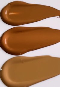 Trois échantillons de maquillage liquide sur fond blanc : marron foncé, marron moyen et beige clair. Chaque échantillon a une texture lisse et brillante.