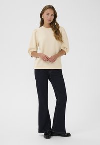 Krämfärgad tröja med puffärmar, i kombination med mörkblå utsvängda jeans. Platta svarta skor fulländar outfiten. Minimalistisk design.