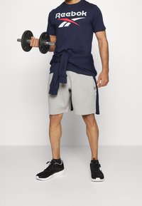 Marinblå Reebok-t-shirt med röd och vit logotyp, grå shorts och svarta träningsskor. Håller en svart hantel i vänster hand.