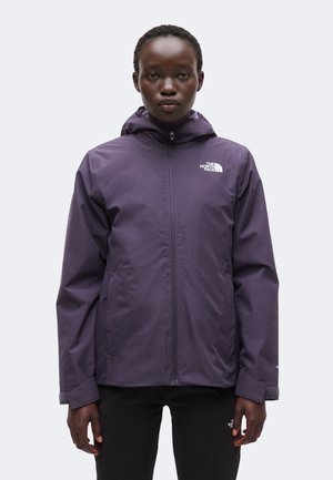 Personne portant une veste zippée violette The North Face avec capuche, debout devant un fond clair uni, regardant vers l’avant.