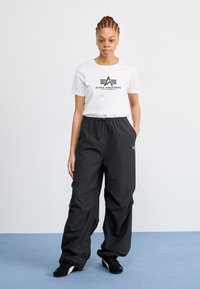 Hvit t-skjorte med svart "Alpha Industries"-logo, kombinert med løstsittende svarte bukser. Buksene har elastisk midje og sidelommer.