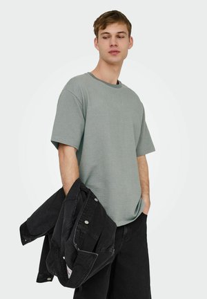 Jonge man draagt een losse grijze textuur korte mouwen overhemd en zwarte broek, houdt een zwarte spijkerjas in zijn linkerhand.