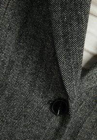 Tessuto grigio a spina di pesce con un motivo testurizzato, dotato di un bottone nero su un capo su misura, che mette in evidenza dettagli netti del revers.