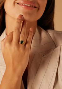 Gold ring featuring an oval green stone surrounded by small clear gems, worn on a hand with a natural nail polish finish.
