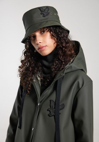 Olivgrüner, wasserdichter Bucket Hat und passender Regenmantel mit schwarzem Adlerlogo und schwarzen Akzenten. Darunter ein gerippter, dunkler Pullover.