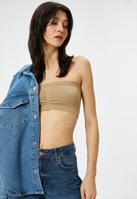 Beżowy top bandeau z teksturowanej tkaniny, noszony pod dżinsową kurtką. Modelka ma na sobie ciemnoniebieskie spodnie z wysokim stanem, podkreślające dopasowaną sylwetkę.