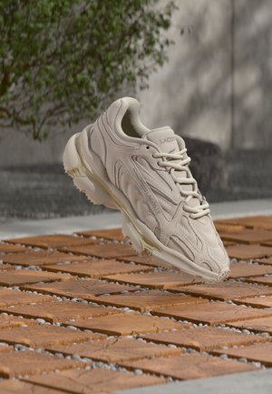 Beige Lacoste sneaker with wavy design and laces suspended above a patterned wooden and pebble ground outdoors.