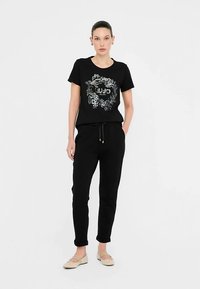 Schwarzes Baumwoll-T-Shirt mit floralem Grafikdesign und dem Schriftzug "JUJO MILANO", kombiniert mit schwarzen Jogginghosen und beigen flachen Schuhen.